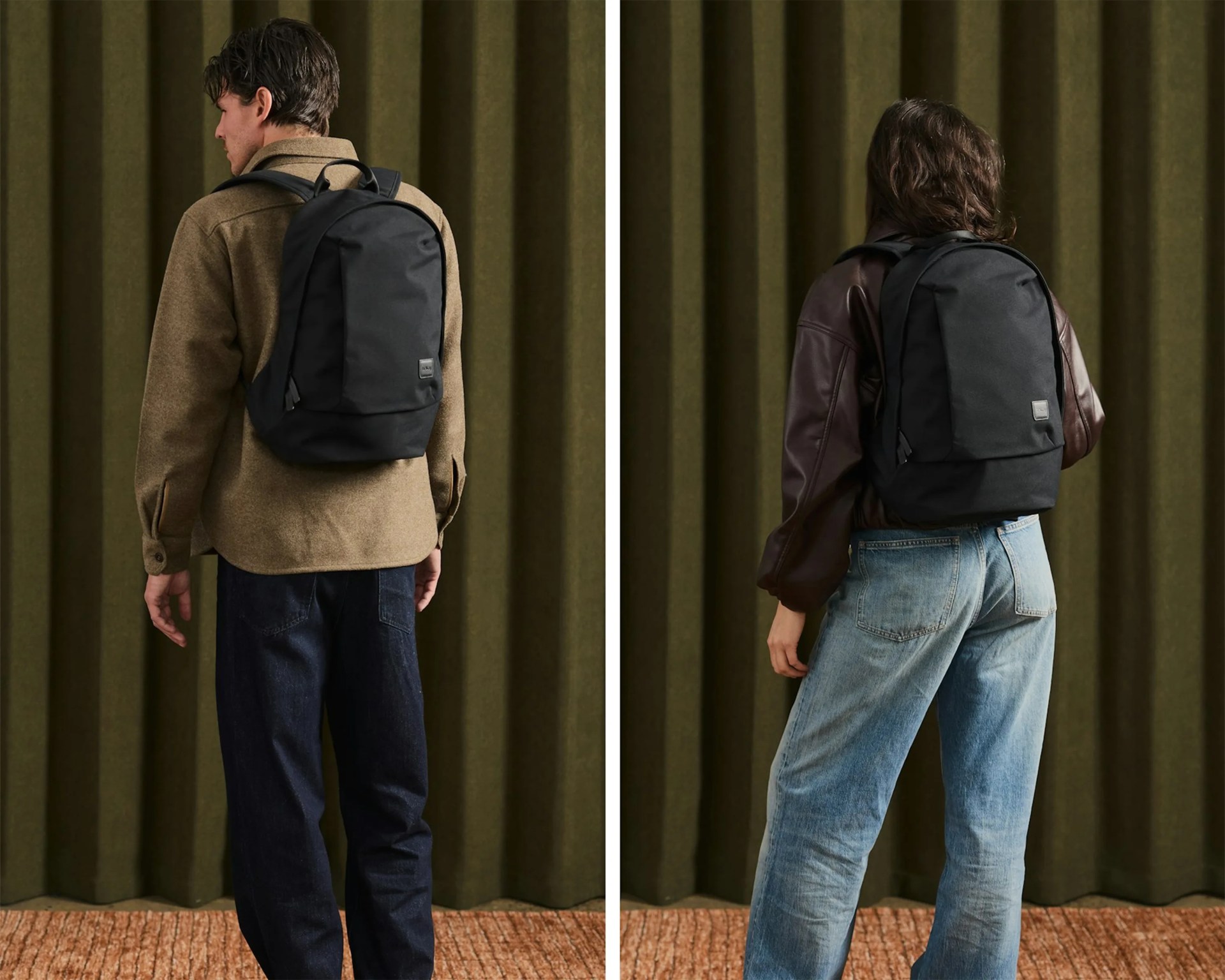 Two people wearing black backpacks, one with a brown jacket and dark jeans, the other with a brown leather jacket and light blue jeans, standing against an olive green curtain.