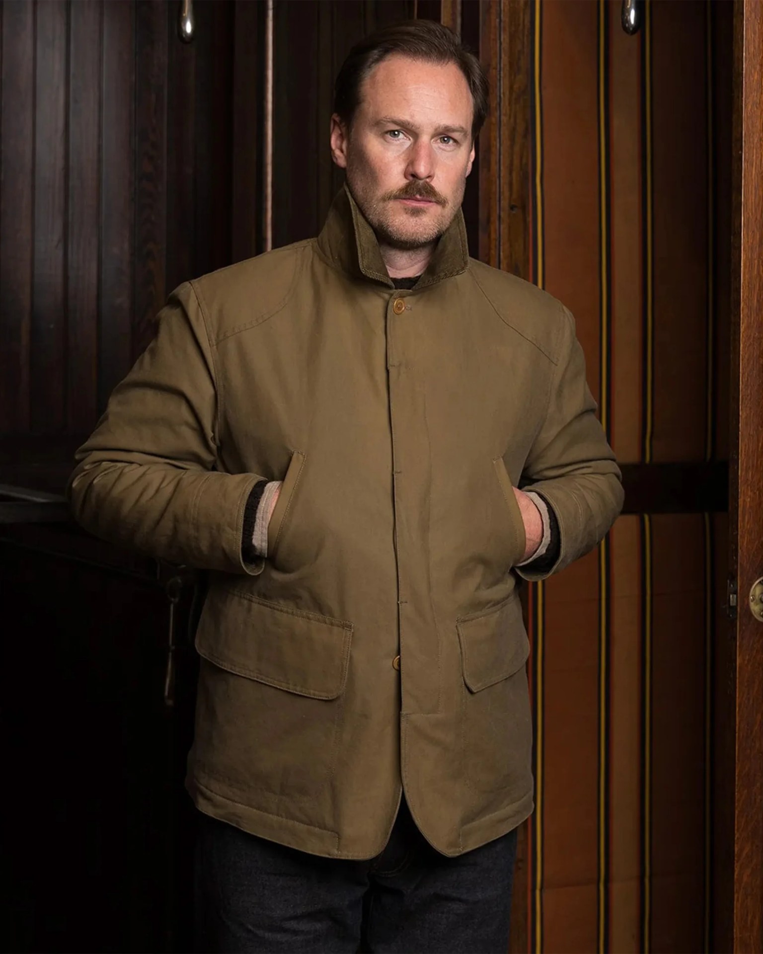 Man wearing a brown buttoned jacket with pockets and a raised collar standing indoors.
