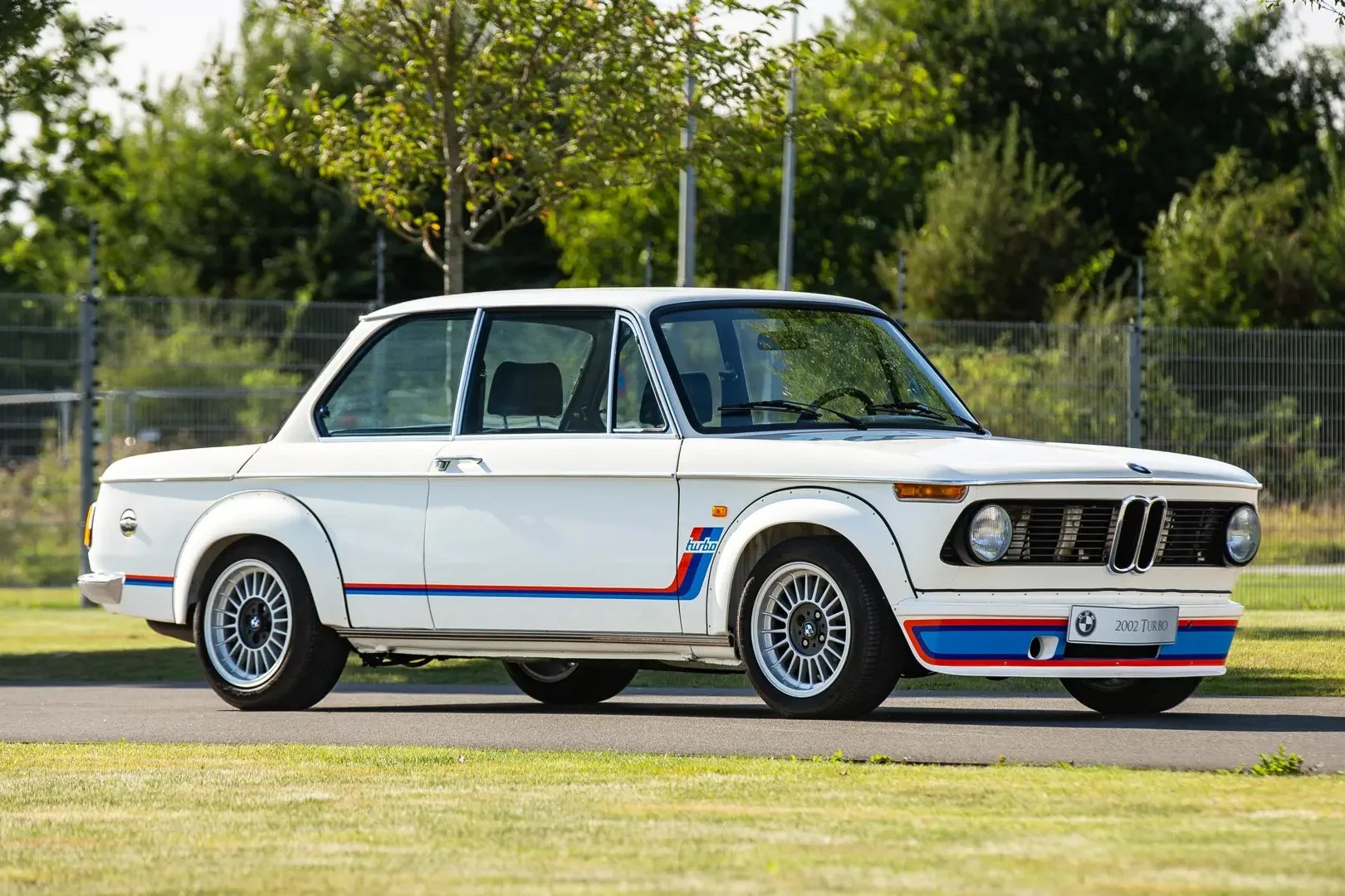White vintage BMW 2002 Turbo with blue and red racing stripes parked on a road with green grass and trees in the background.