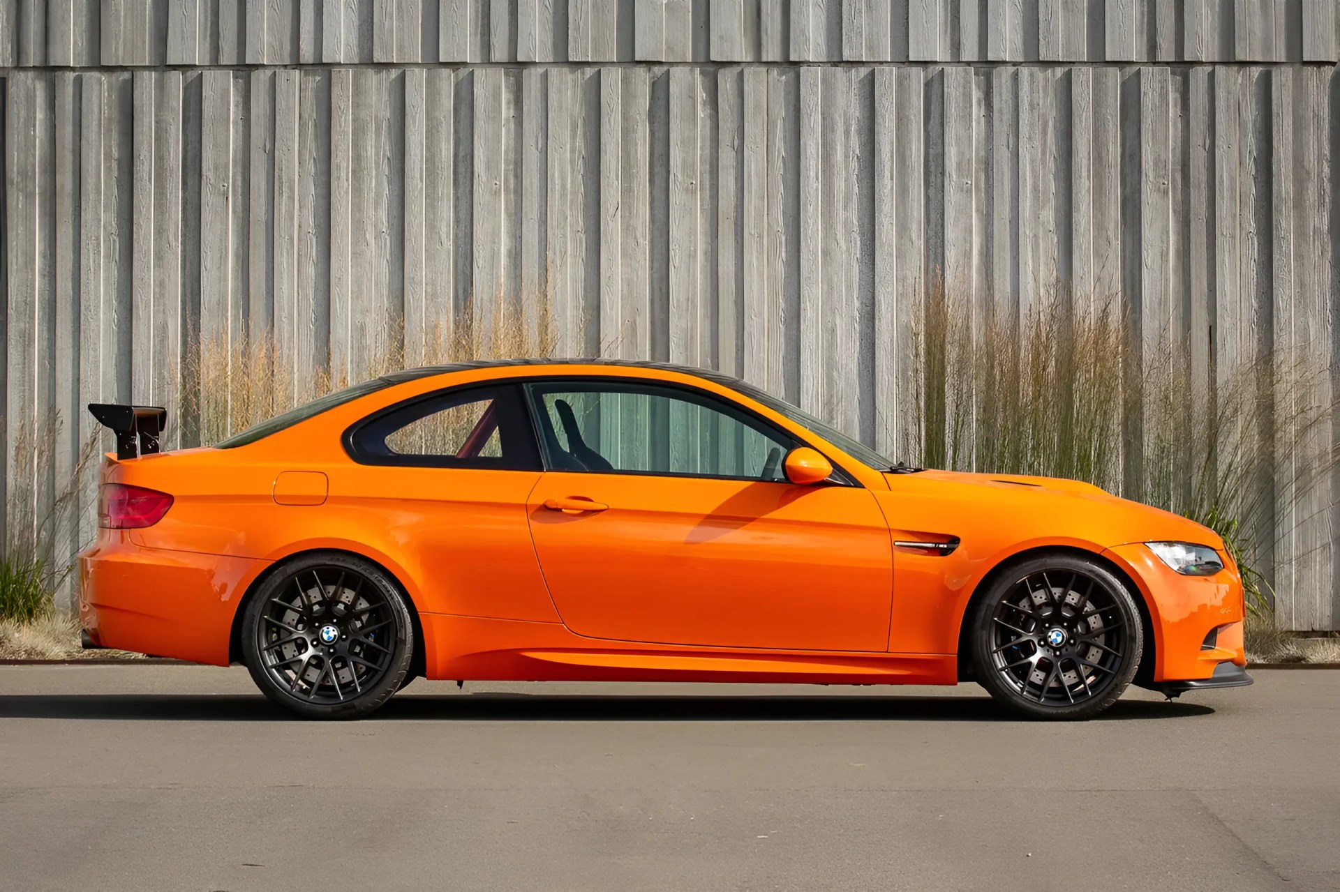 Bright orange BMW coupe with black wheels and a large rear spoiler parked in front of a wooden wall.