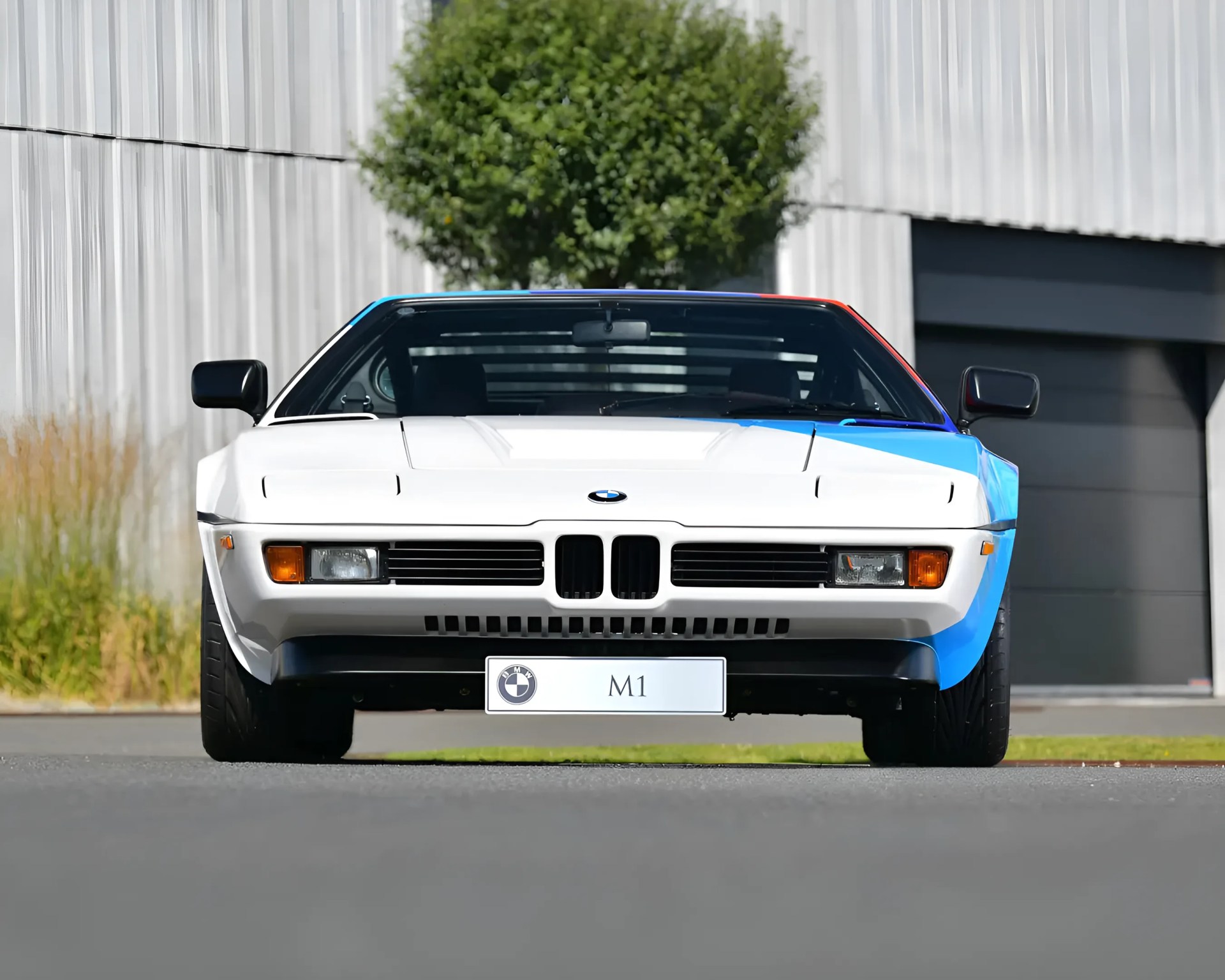 Front view of a white and blue BMW M1 sports car parked on a road with a building and tree in the background.
