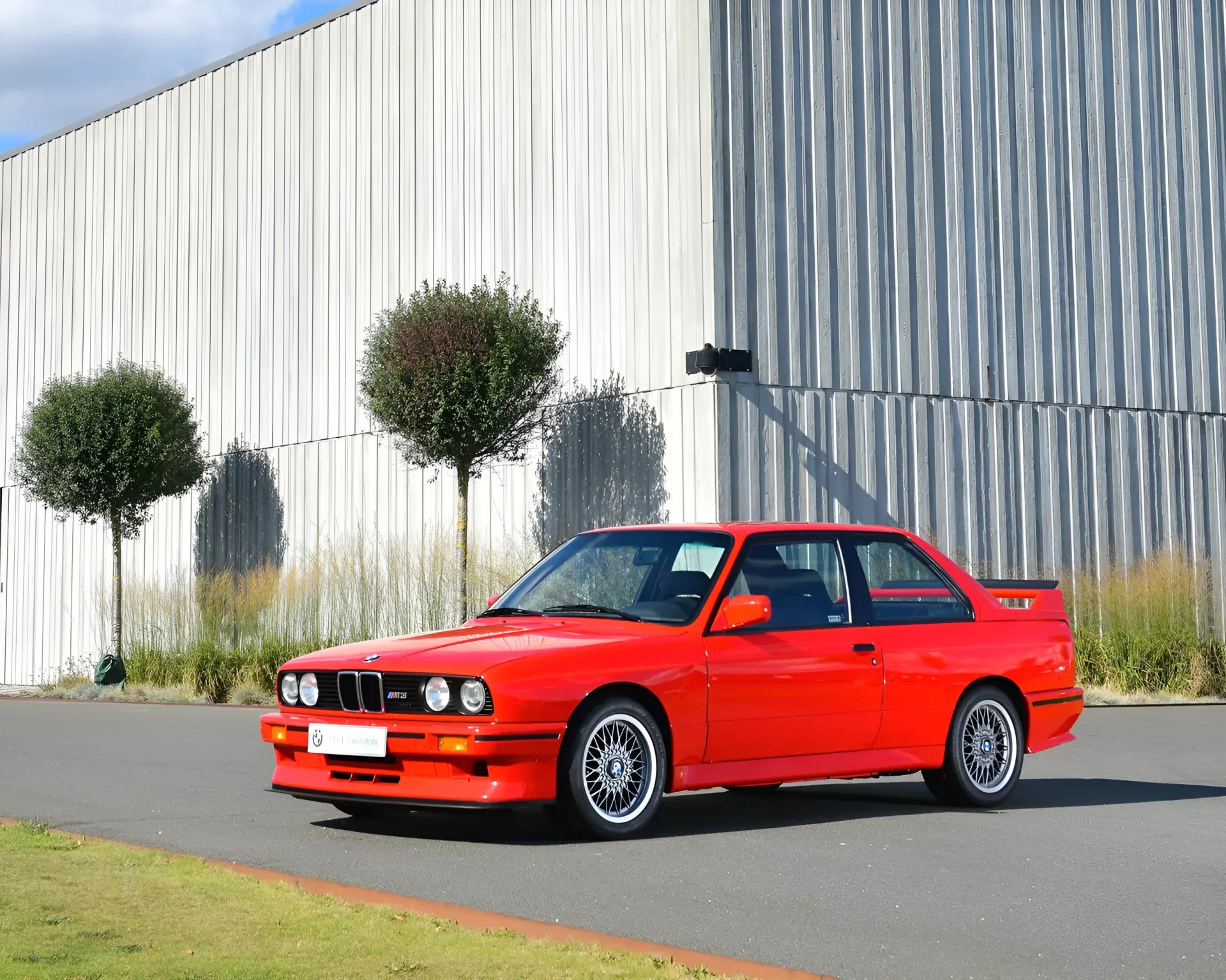Red BMW E30 M3 coupe with mesh alloy wheels parked on a paved area near a white industrial building.