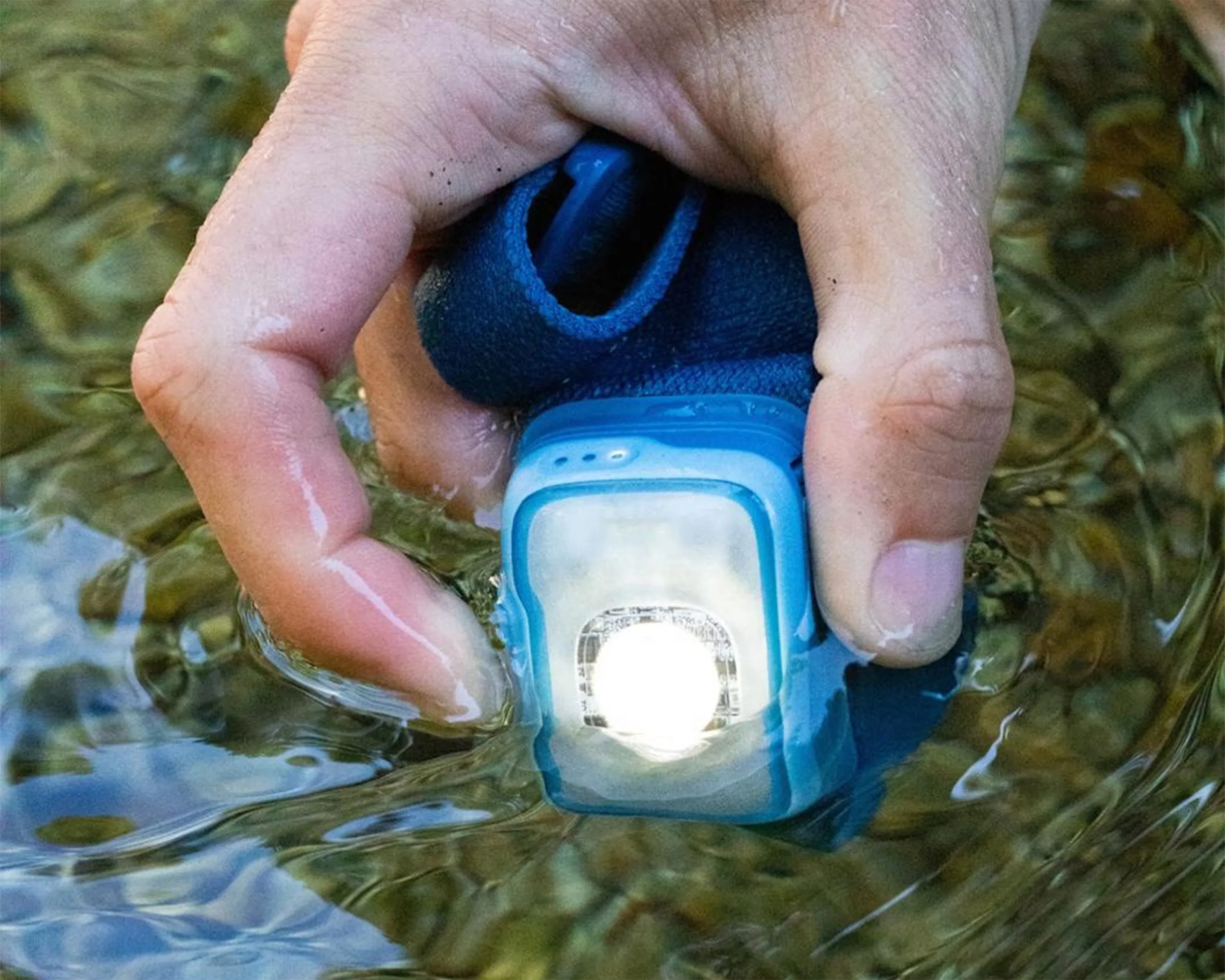 Hand holding a blue waterproof headlamp submerged in clear water.