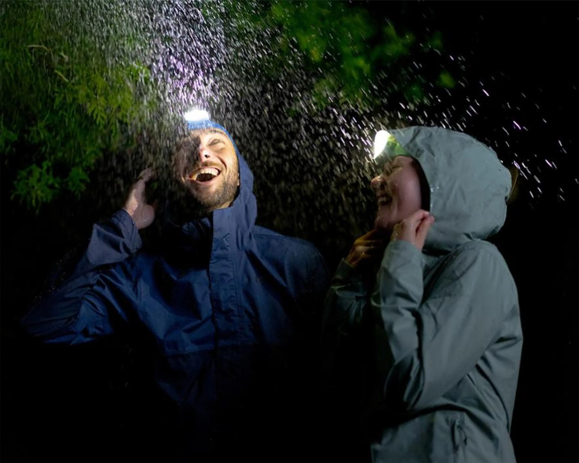 Two people wearing rain jackets and headlamps laughing under falling rain at night.