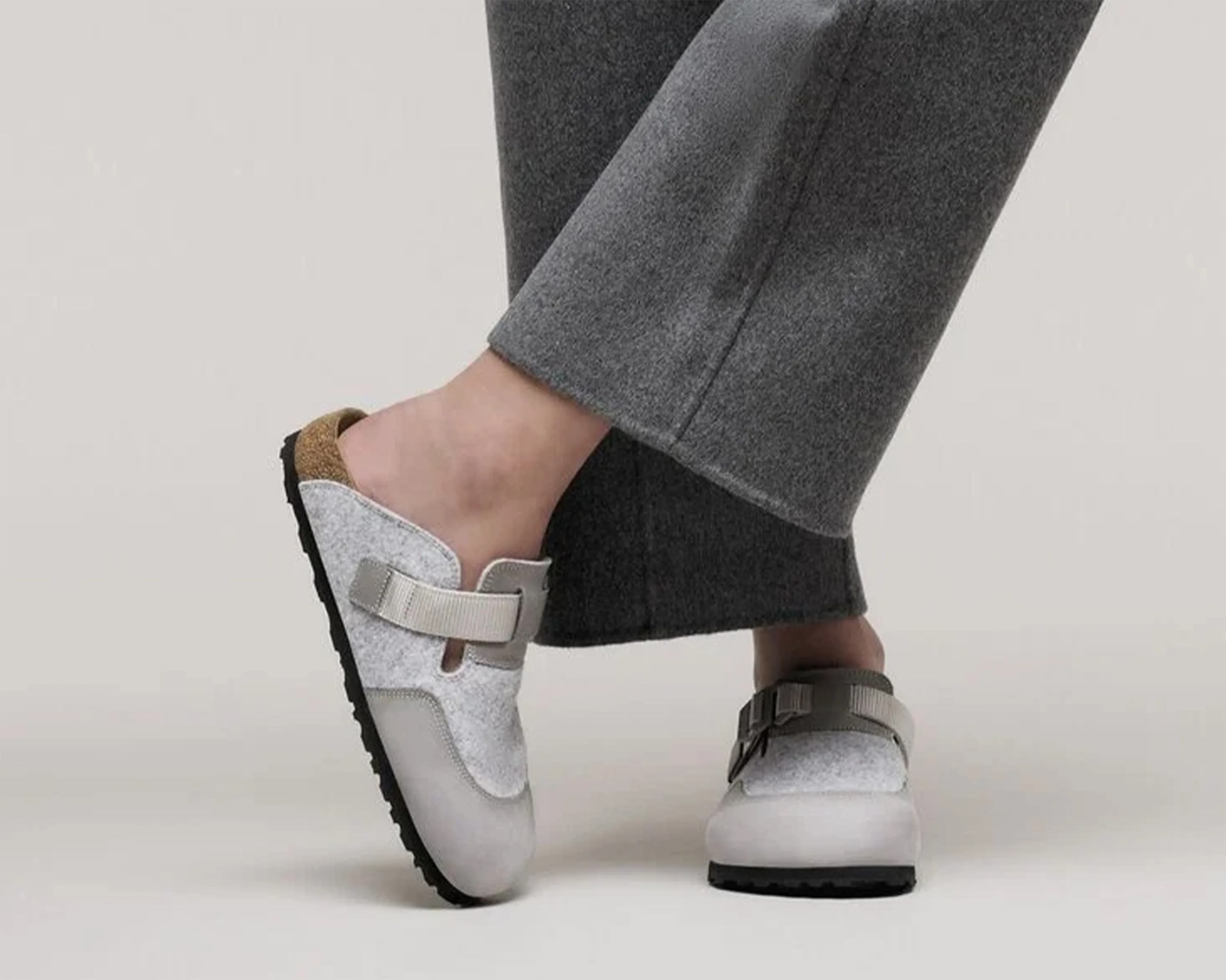 Pair of light gray slip-on clogs with adjustable straps, cork footbed, and black soles worn with gray pants.