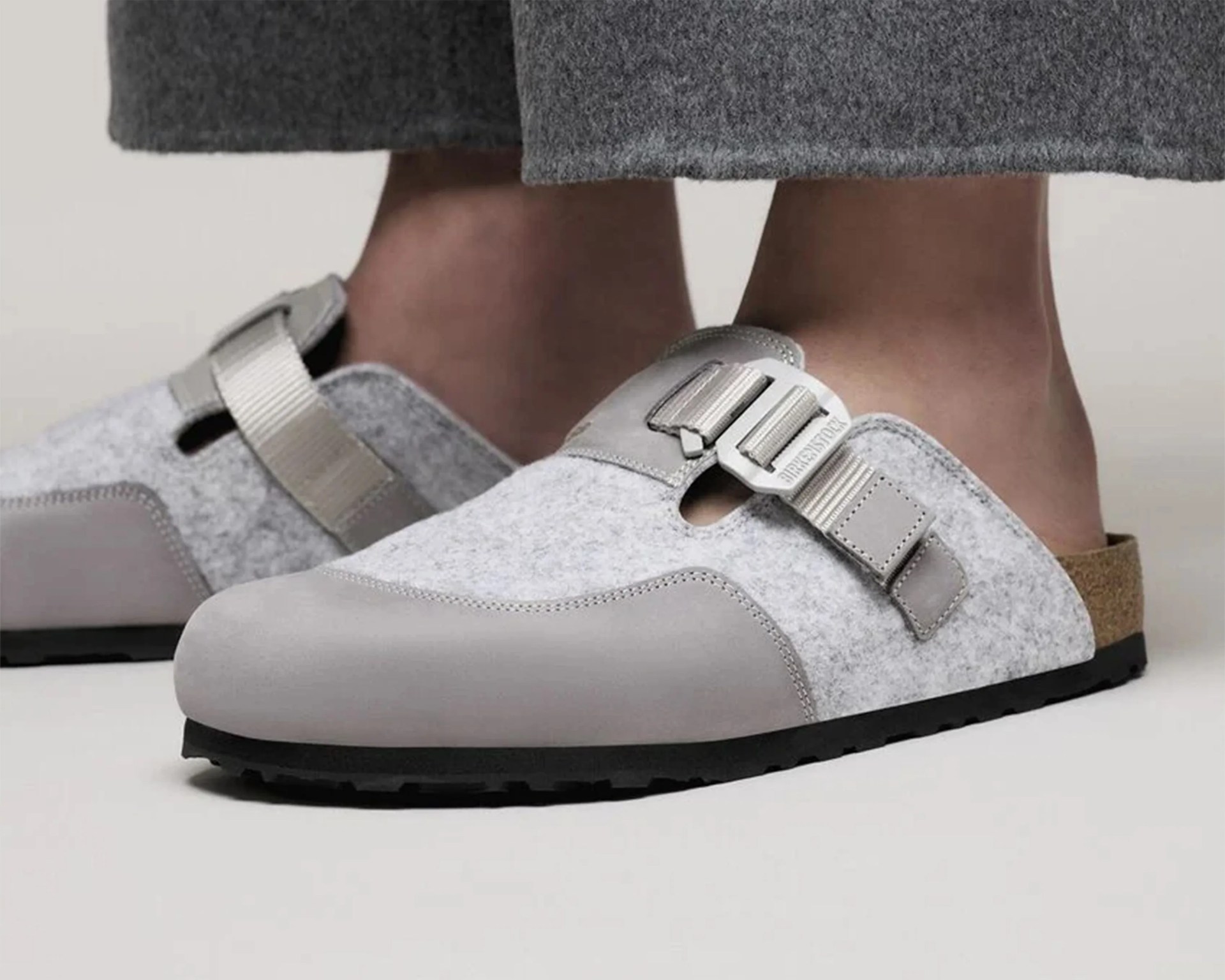 Gray felt and leather slip-on clogs with adjustable straps and black soles worn with gray pants.