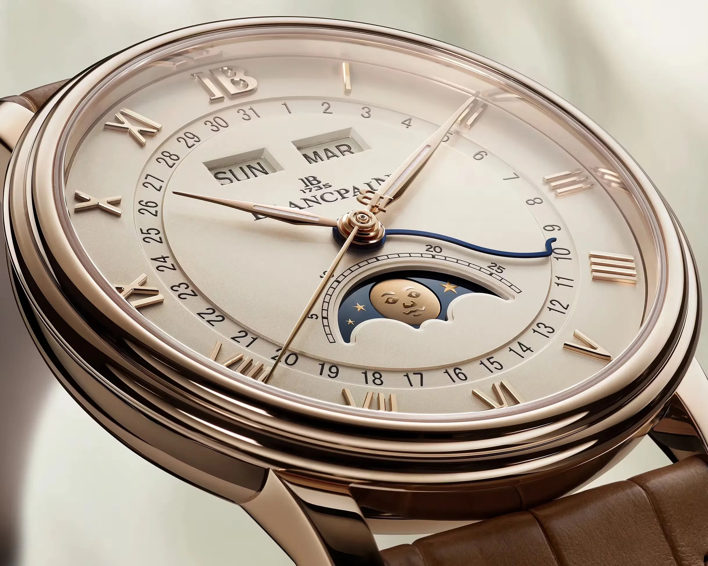 Close-up of a rose gold wristwatch with Roman numerals, day and month windows, and a moon phase indicator on a beige dial.
