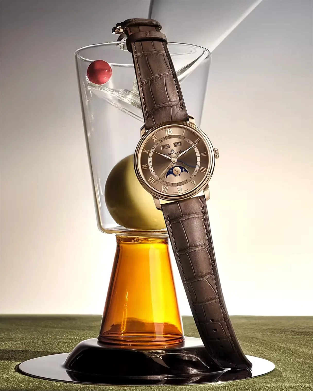 Gold-tone wristwatch with brown leather strap and moon phase dial displayed on an amber and clear glass sculpture.
