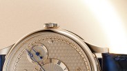 Close-up of a gold watch face with intricate textured dial, blue hands, Roman numerals, and a black leather strap.