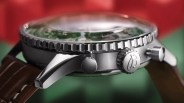 Close-up of a stainless steel watch case with a ridged bezel, crown engraved with a "B," and brown leather strap.