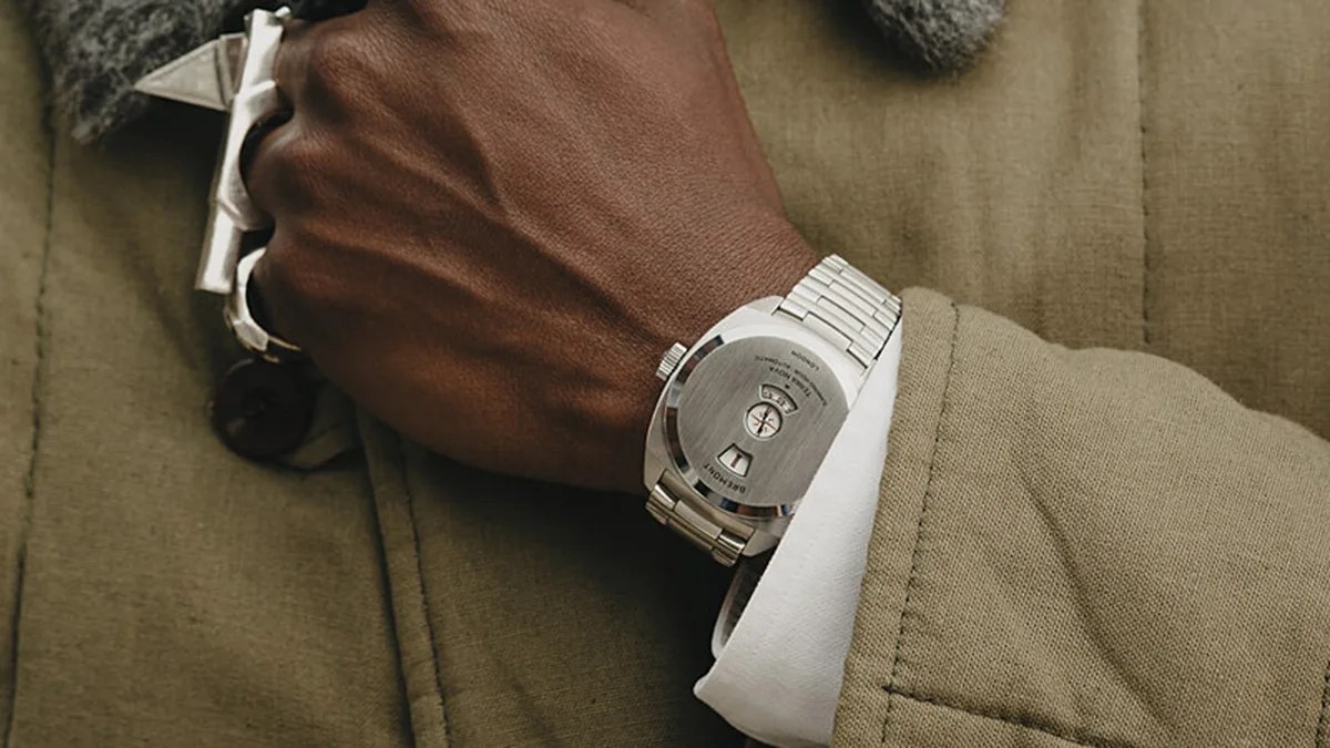 Silver metal wristwatch with a brushed back case and a metal link bracelet worn on a wrist over a white shirt cuff and olive green jacket.