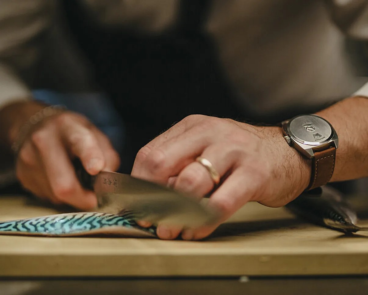 Hands slicing a fish fillet with a knife on a wooden cutting board, one hand wearing a watch with a brown leather strap.