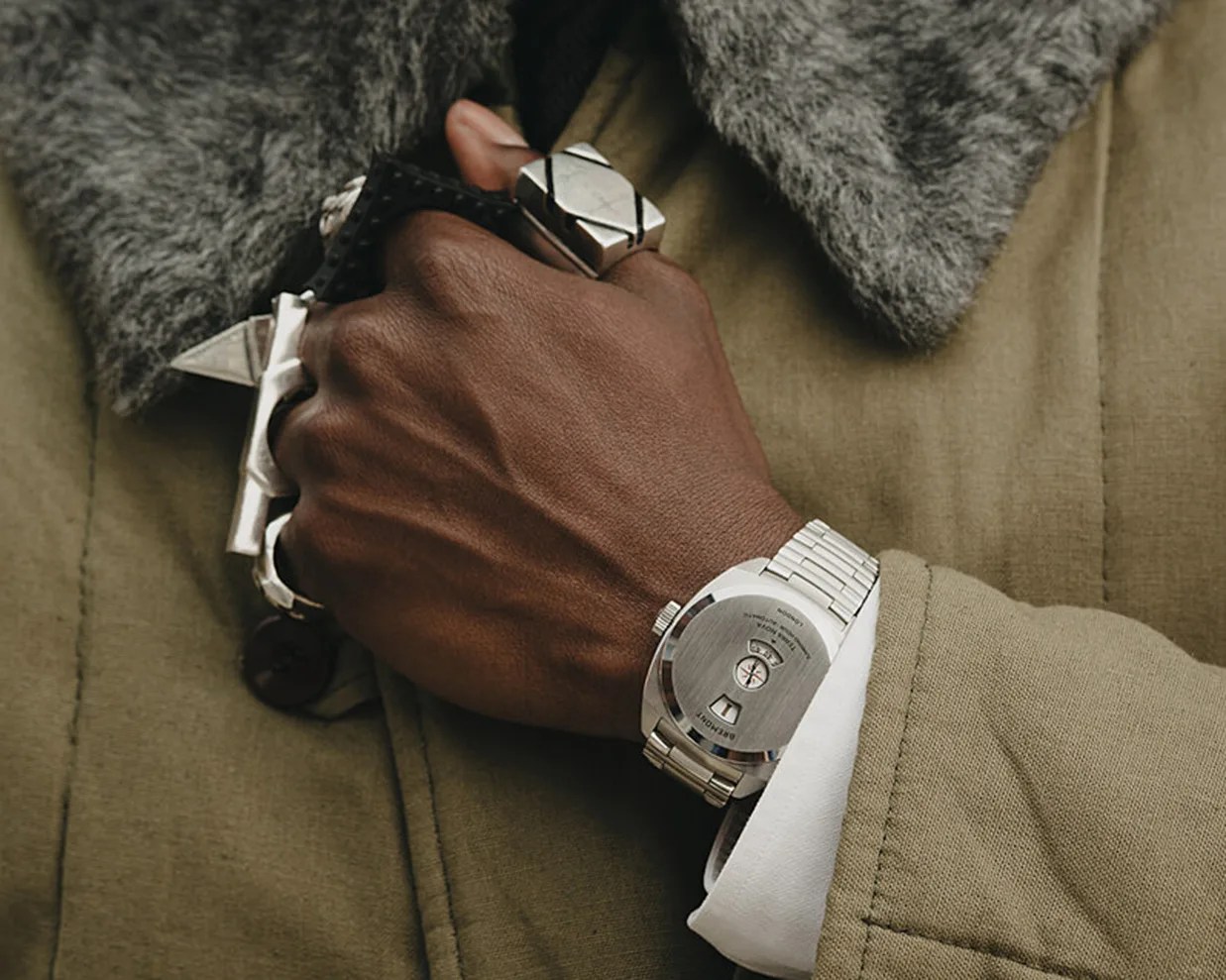 Close-up of a wrist wearing a silver metal watch with a brushed back, a white cuff, and a green coat with a gray fur collar.