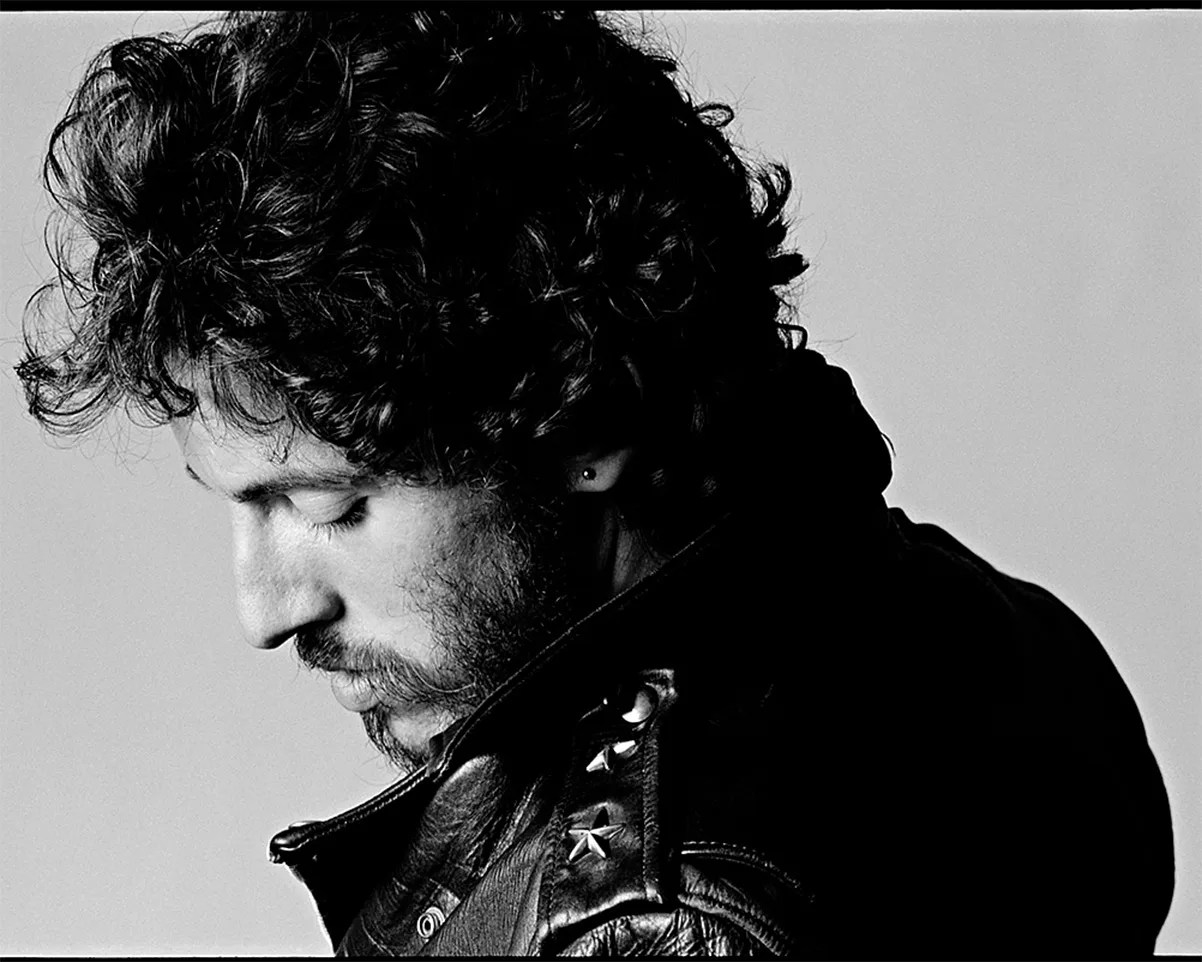 Black and white close-up of a person with curly hair and a beard wearing a leather jacket with star studs, looking down.