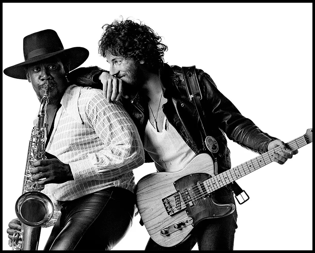 Black-and-white photo of a man playing saxophone wearing a wide-brim hat and another man with curly hair playing electric guitar in a leather jacket.