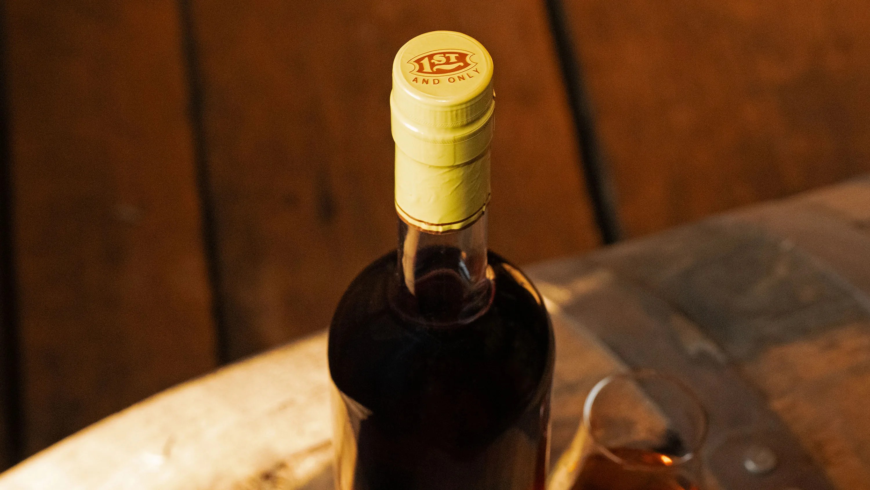 Dark glass bottle with a yellow cap labeled "1st and only" on a wooden surface next to a glass with amber liquid.