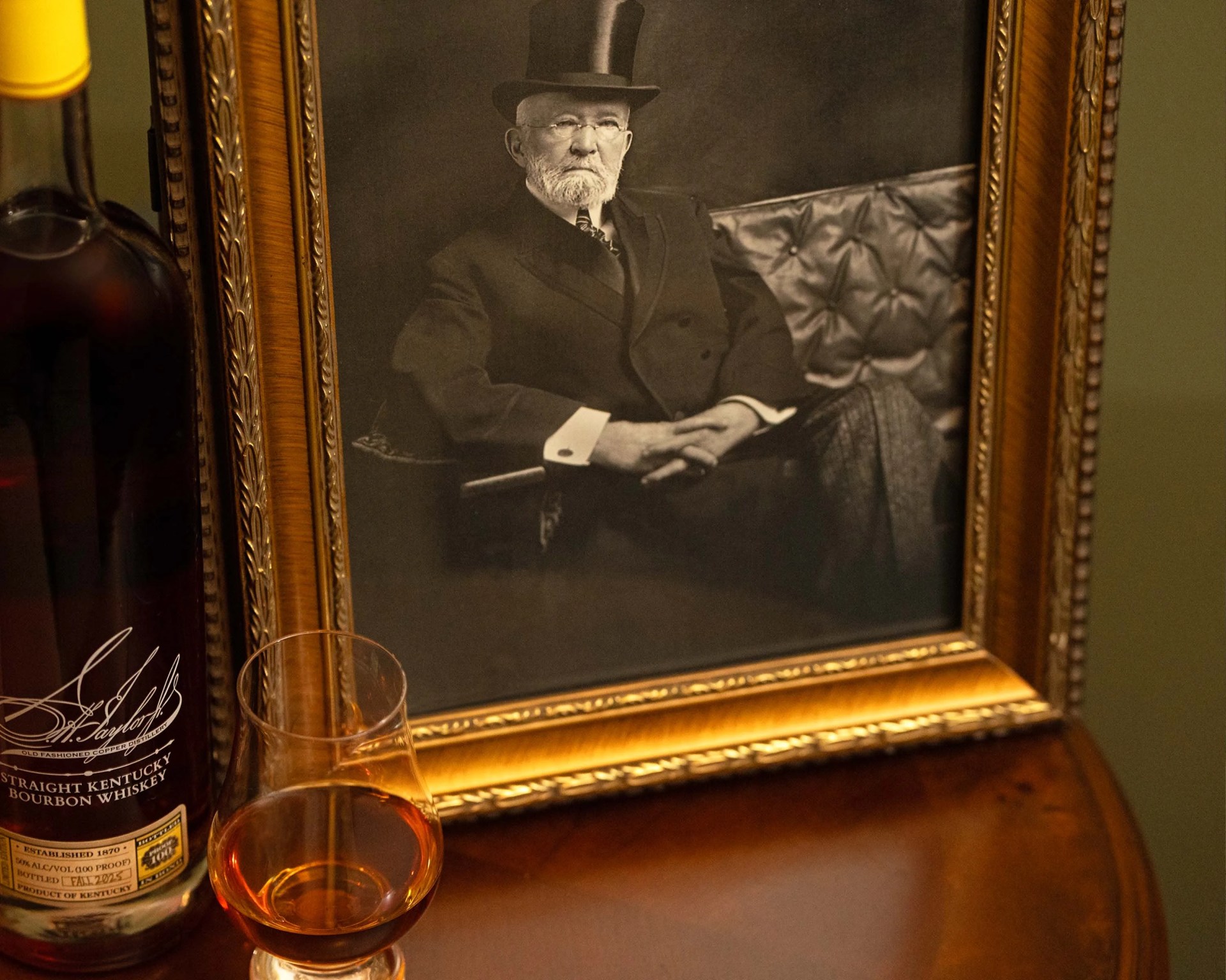 Glass of amber bourbon whiskey next to a bottle and a framed black-and-white portrait of a man in a top hat.