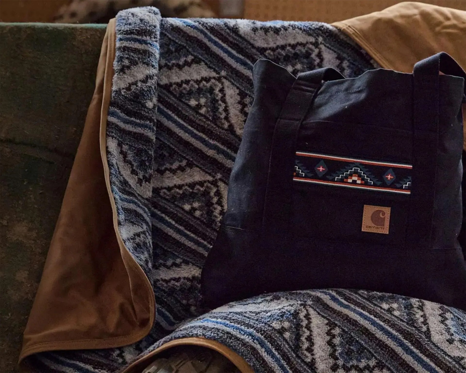 Black Carhartt tote bag with geometric patterned stripe and leather logo patch on a blue and gray patterned blanket with brown trim.