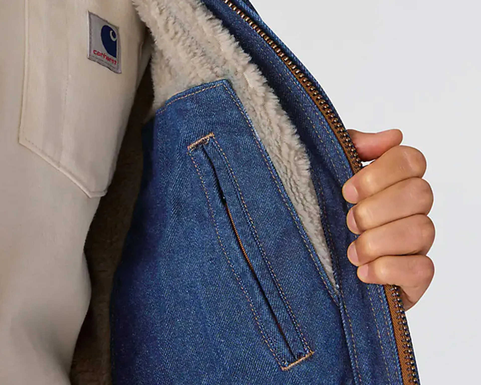 Hand holding open a blue denim jacket with beige sherpa lining and a visible Carhartt logo on a beige shirt.