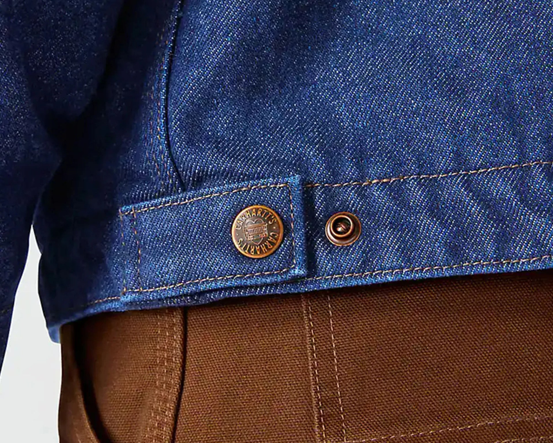 Close-up of blue denim jacket hem with copper Carhartt button over brown pants.