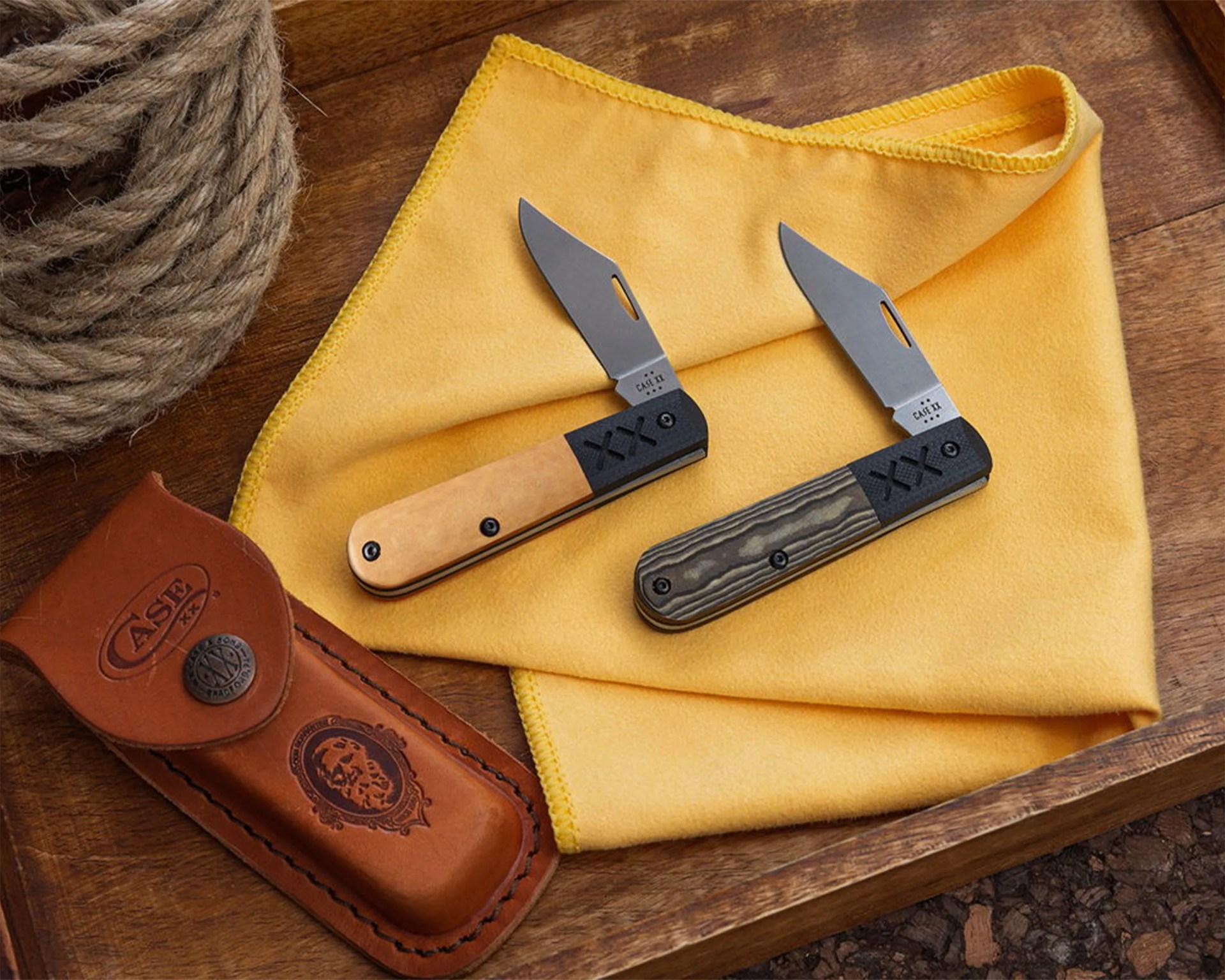 Two folding pocket knives with black and tan handles on a yellow cloth next to a brown leather sheath embossed with "CASE XX".
