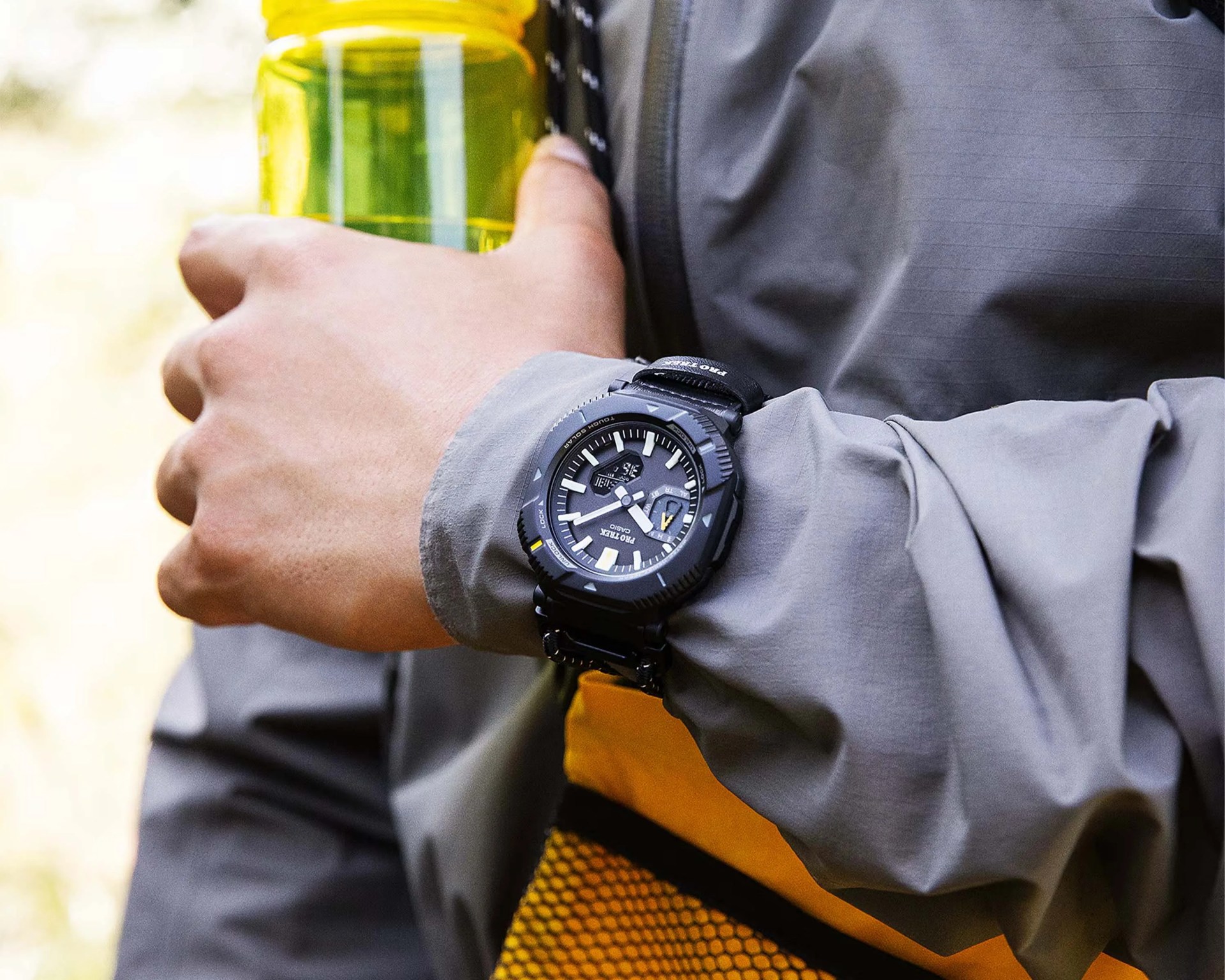 Black Casio Pro Trek digital-analog watch with a black resin strap worn on a wrist holding a yellow water bottle.