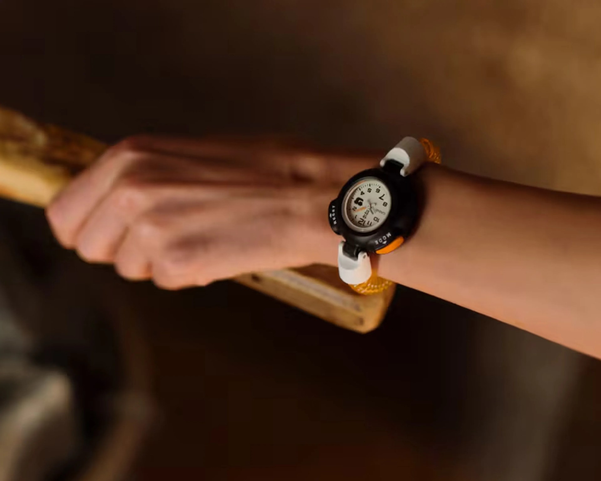 Wristwatch with a white dial, black bezel, and orange and white strap worn on a wrist holding a wooden object.