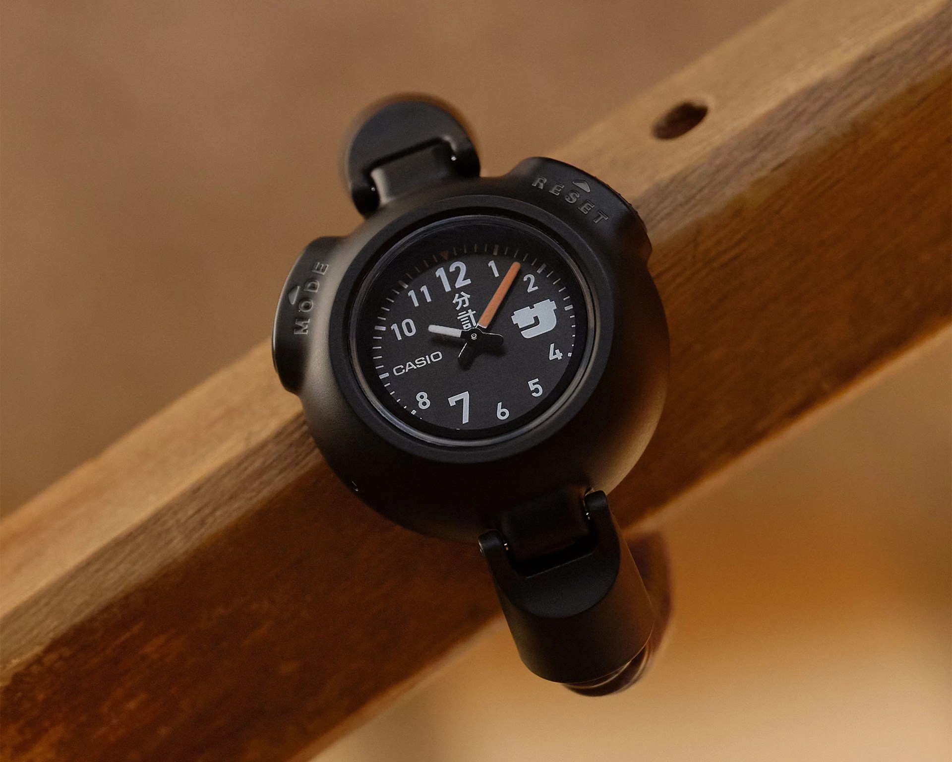 Black Casio wristwatch with round face, white numerals, and orange minute hand on a wooden surface.