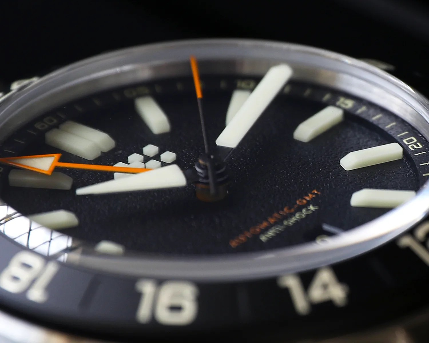 Close-up of a black watch face with large white hour markers, orange-tipped hands, and a date display showing the number 16.
