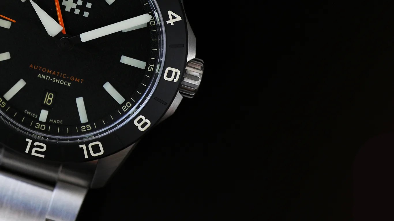 Close-up of a black dial automatic GMT watch with white hour markers, orange second hand, and stainless steel case and bracelet.