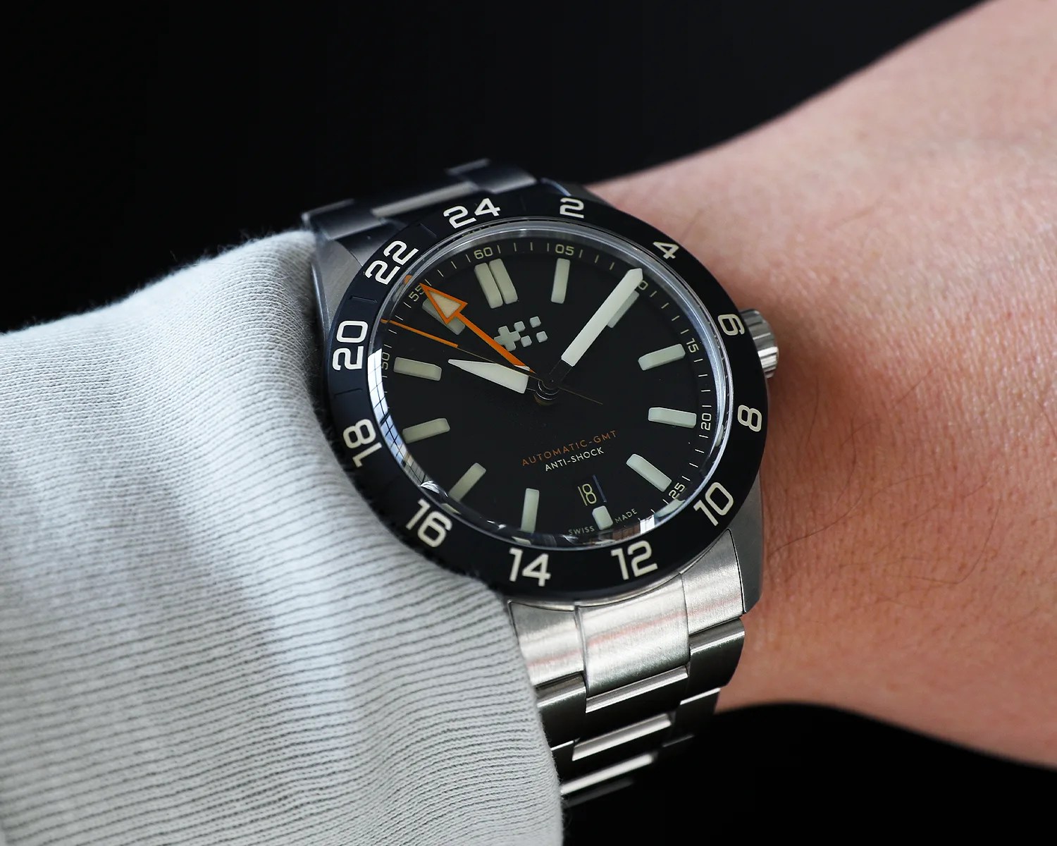 Black dial wristwatch with white hour markers, orange GMT hand, and stainless steel bracelet worn on a wrist.