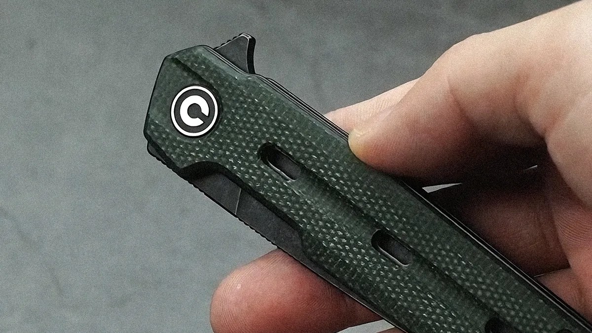Hand holding a closed folding knife with a textured dark green handle and a circular logo near the pivot.