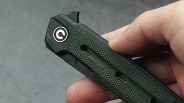 Hand holding a closed folding knife with a textured dark green handle and a circular logo near the pivot.