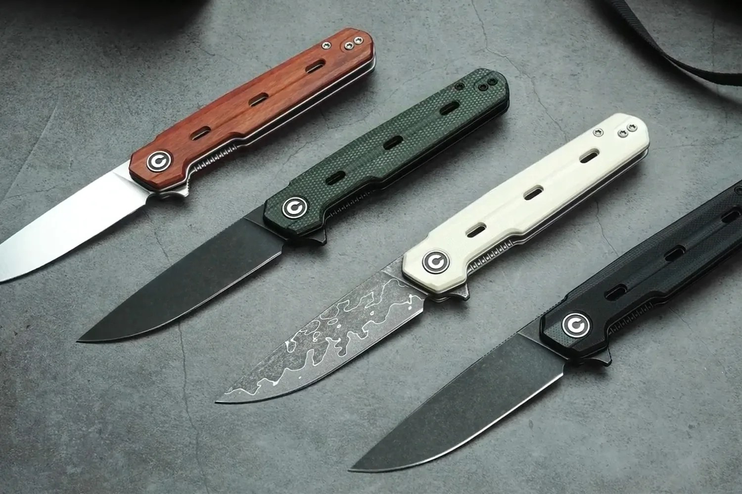 Four folding knives with different handle colors&mdash;wood, green, white, and black&mdash;laid out on a gray surface.