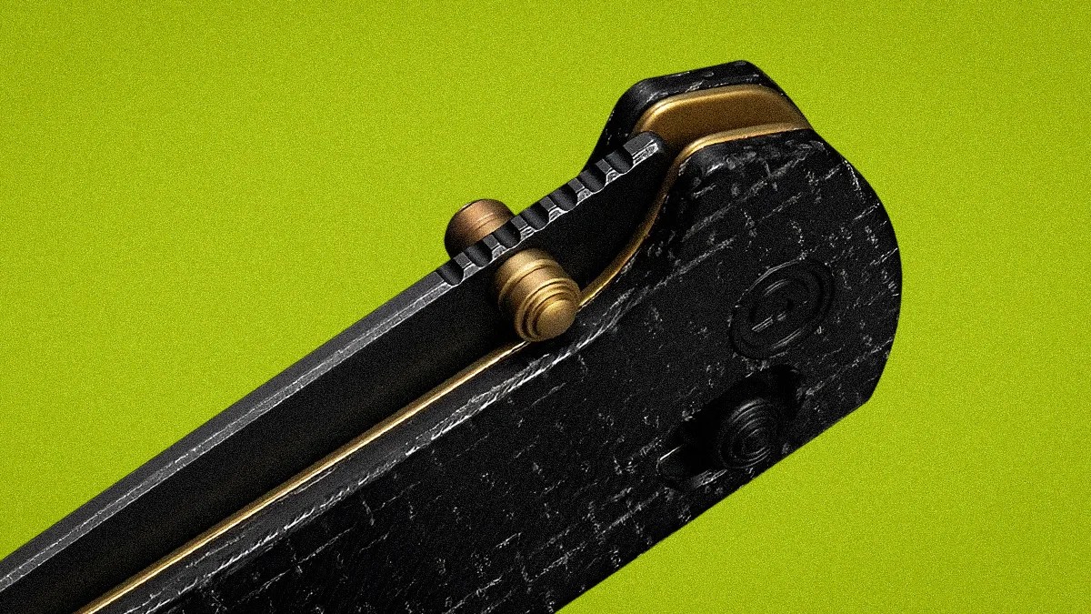 Close-up of a black textured folding knife handle with brass accents and a thumb stud on a green background.