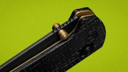 Close-up of a black textured folding knife handle with brass accents and a thumb stud on a green background.