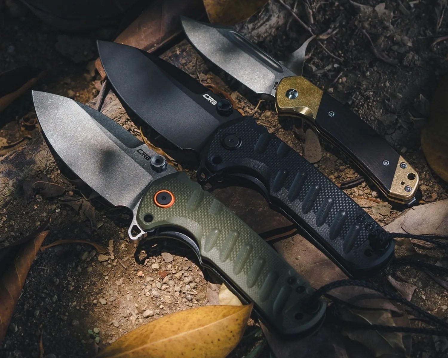 Three folding knives with textured handles in black, green, and black with brass accents, laid on dirt and leaves.