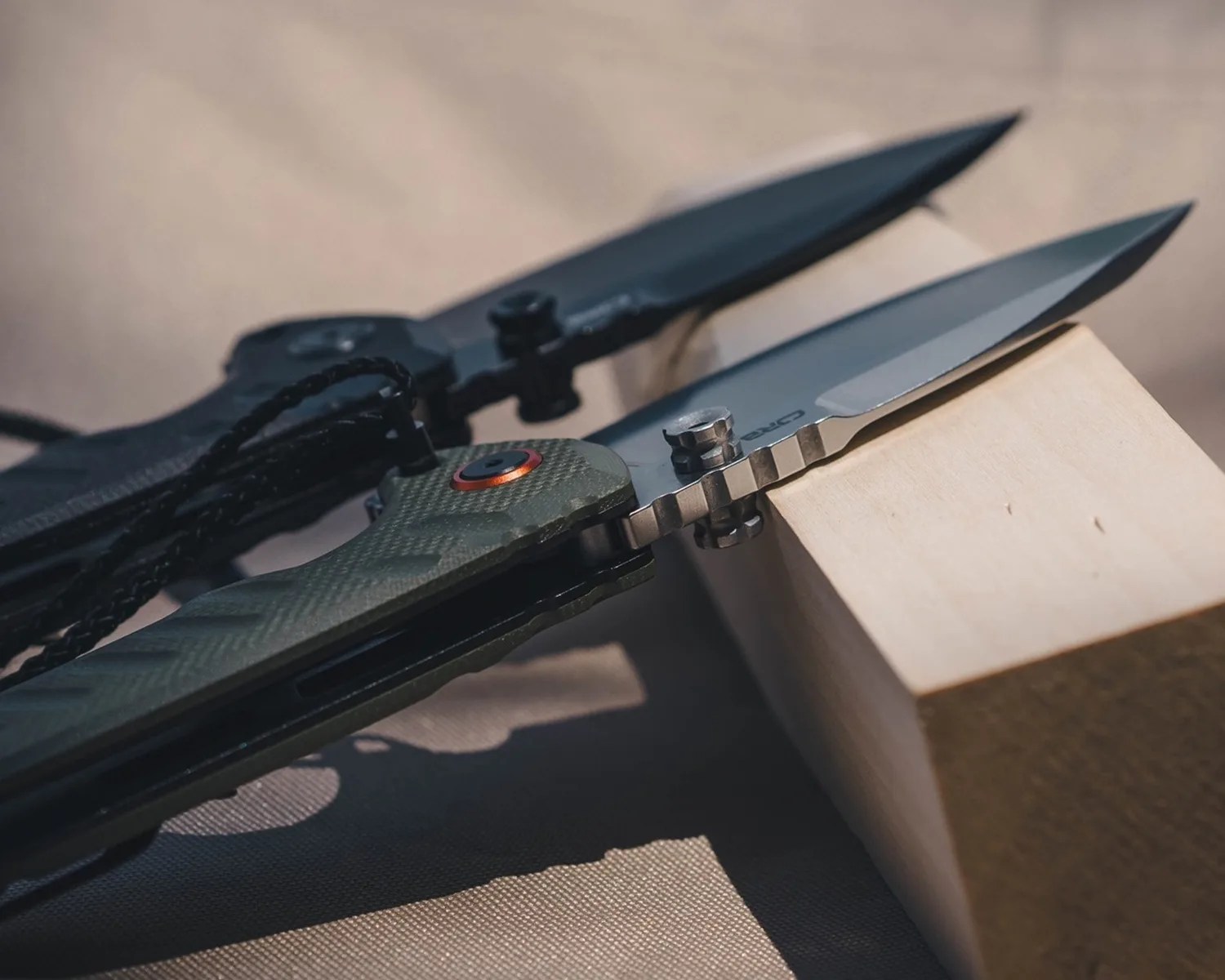 Two folding knives with partially serrated blades resting on a wooden block, one with a green textured handle and the other with a black handle.