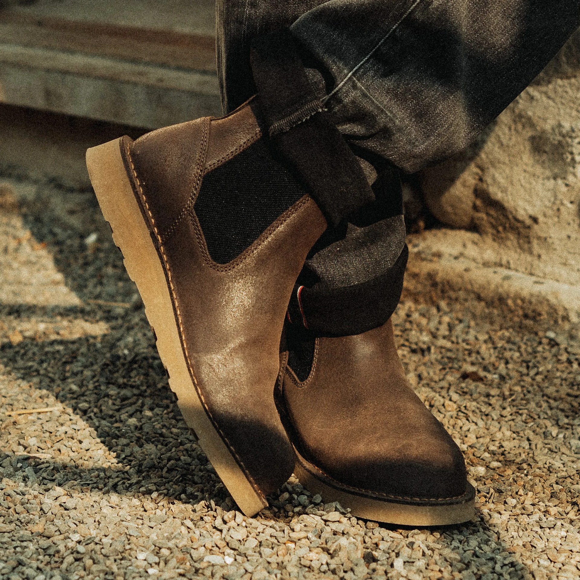Brown leather Chelsea boots with black elastic side panels and light tan soles worn with cuffed dark jeans on gravel.