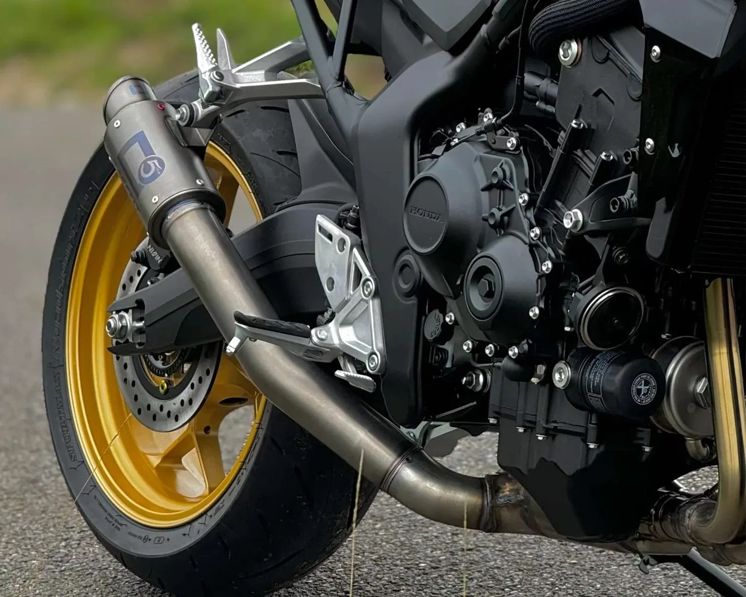 Close-up of a motorcycle's rear wheel with gold rim, black tire, and a silver exhaust pipe attached to a black engine.