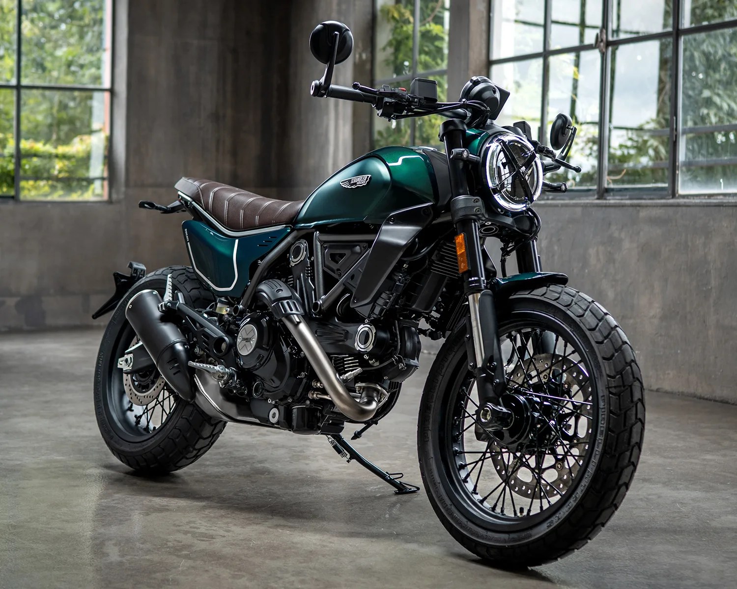 Dark green and black scrambler motorcycle with brown leather seat parked indoors on a concrete floor.