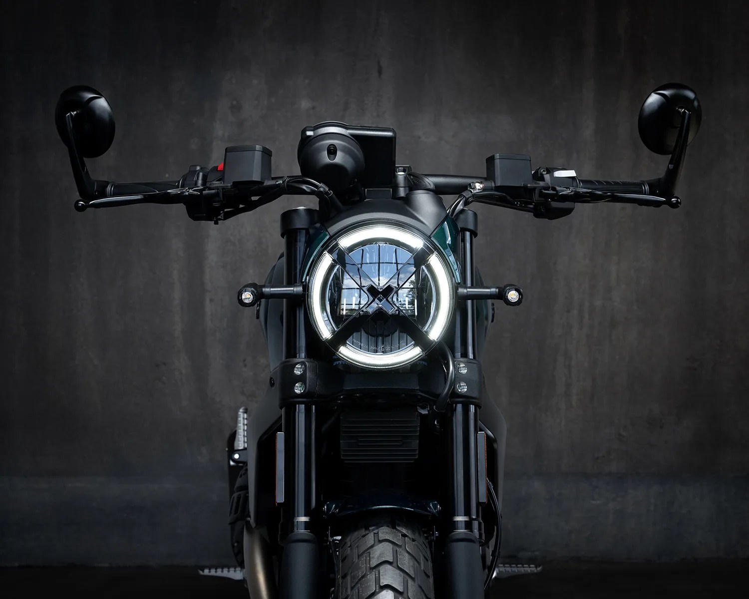 Front view of a black motorcycle with a circular LED headlight and black handlebars against a dark gray background.