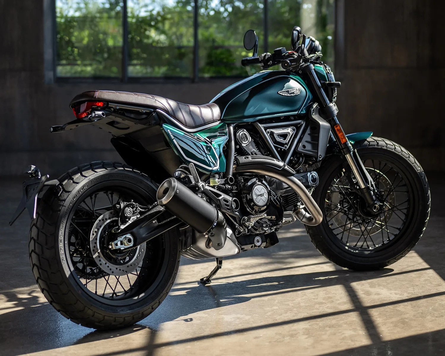Teal Ducati Scrambler motorcycle with black exhaust and brown leather seat in a sunlit garage.