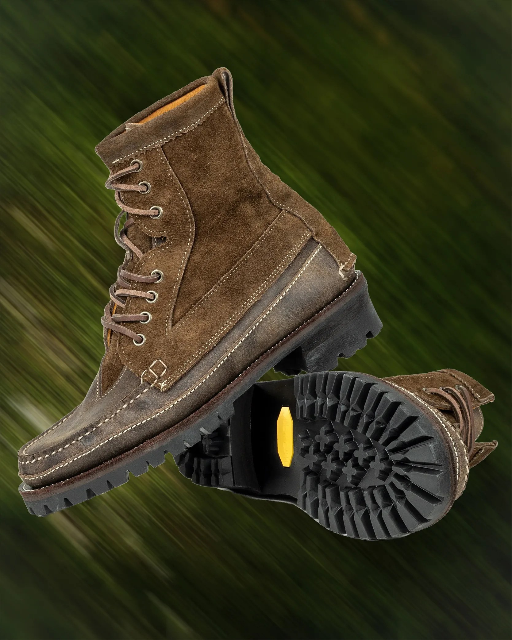Pair of brown suede and leather lace-up boots with black lug soles against a blurred green background.