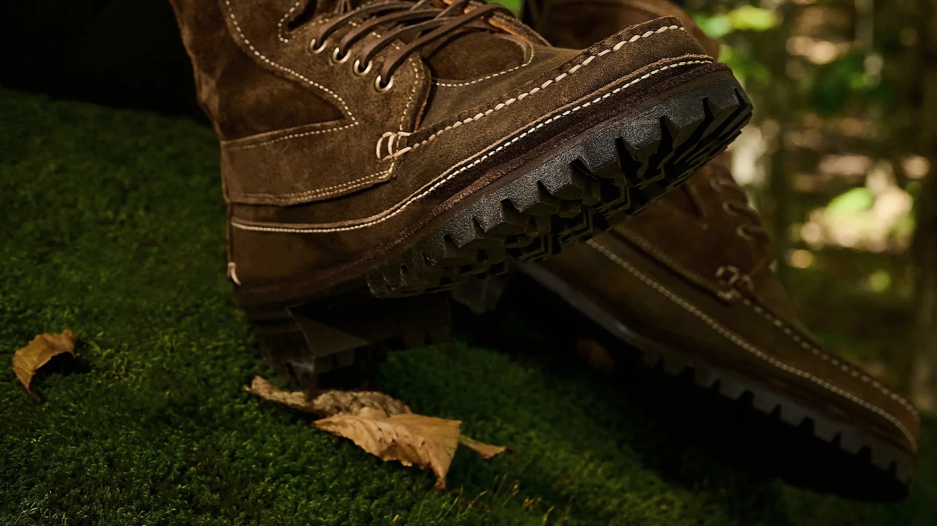 Brown suede hiking boots with thick black lug soles on green moss with dry leaves.