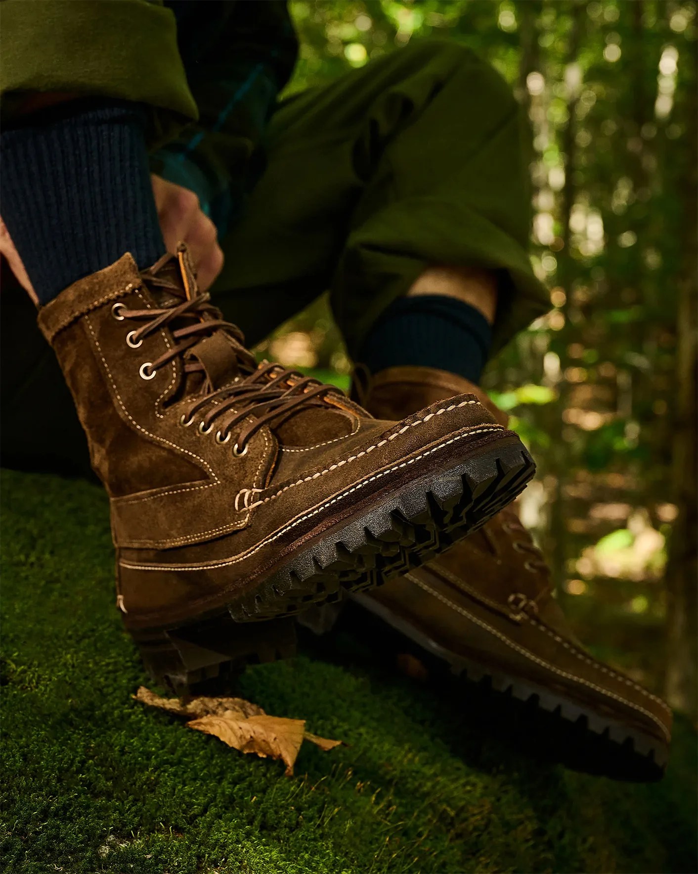 Brown suede lace-up boots with thick black lug soles worn by a person sitting on mossy ground.