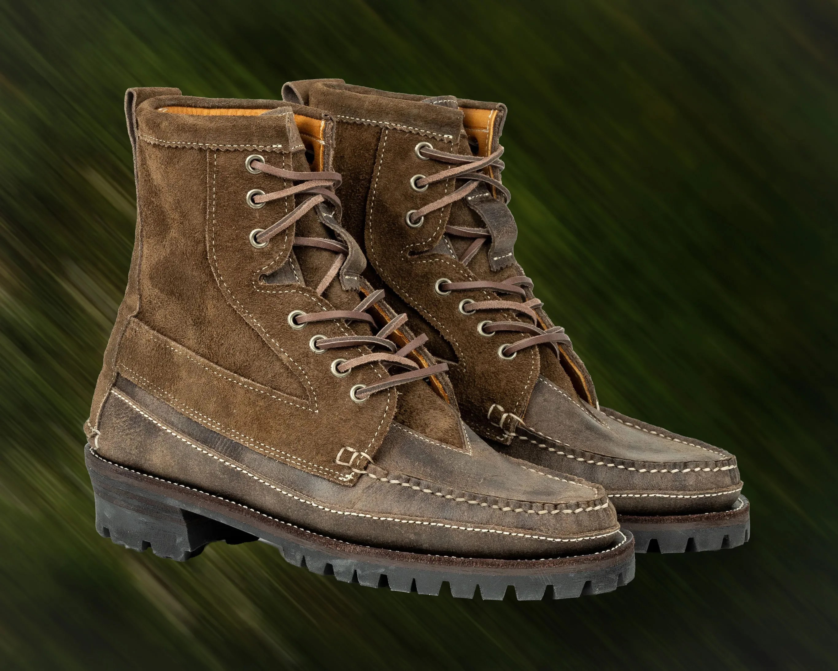 Pair of brown suede and leather lace-up boots with rugged black soles on a blurred green background.