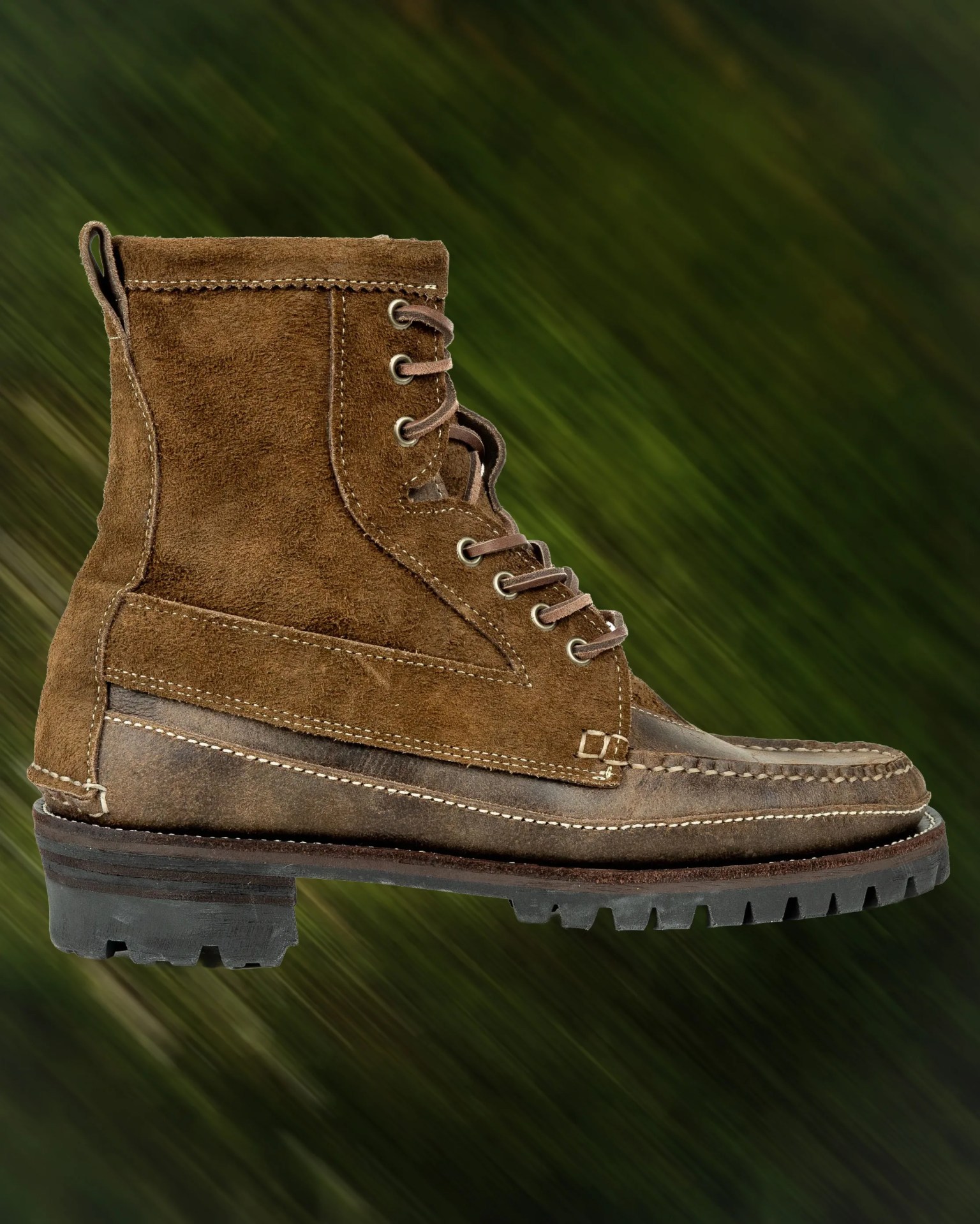 Brown suede and leather lace-up boot with a rugged black sole against a blurred green background.