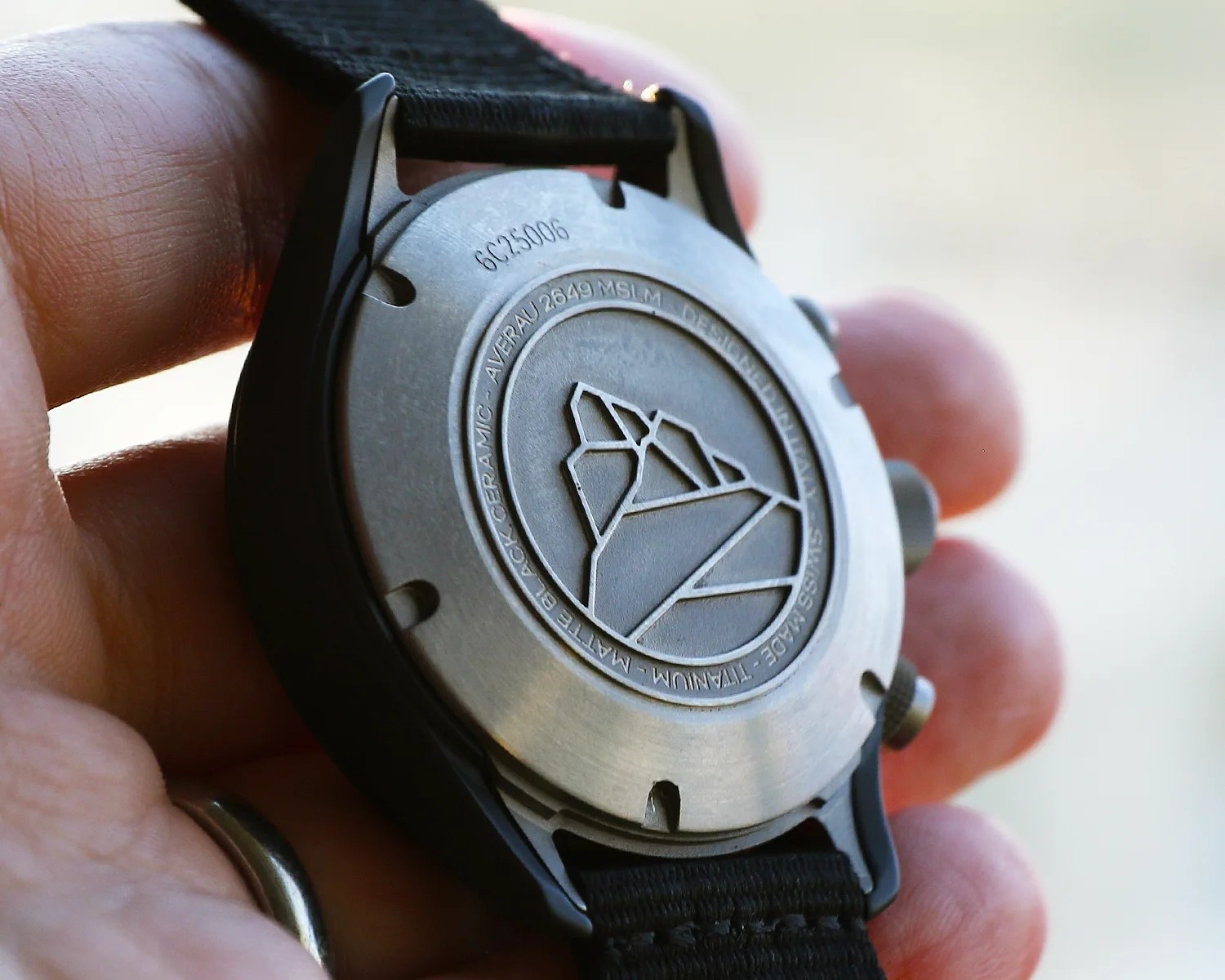 Close-up of a titanium watch back with engraved geometric design and black fabric strap held in a hand.