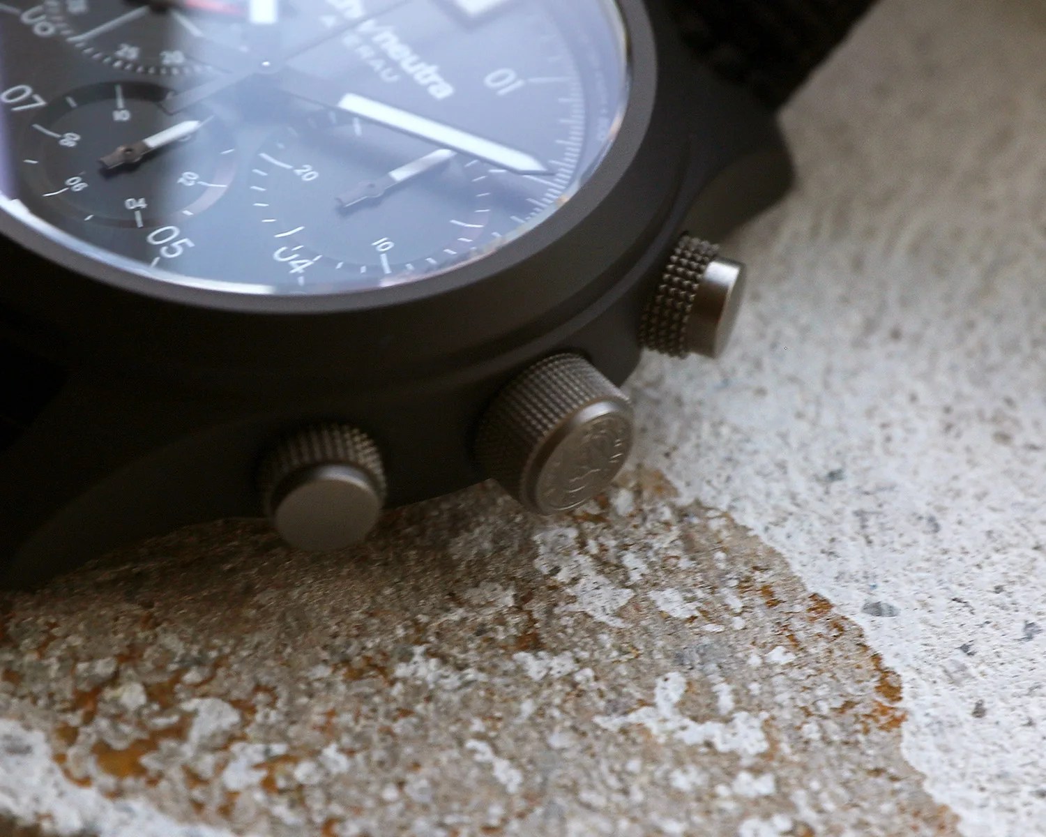 Close-up of a black wristwatch with three textured metal crowns on a rough, speckled stone surface.